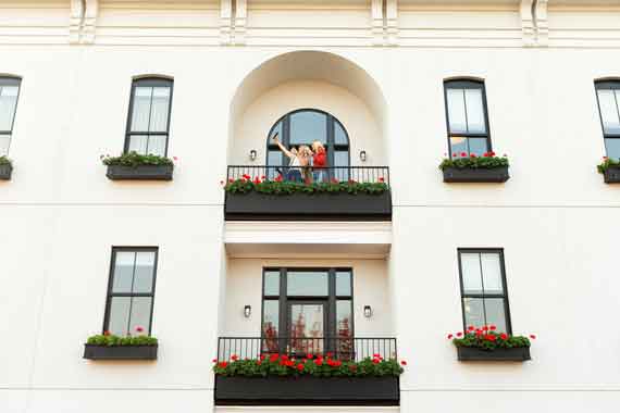 White multi-story building with flower boxes on each balcony; a couple stands on the upper central balcony overlooking the scene.