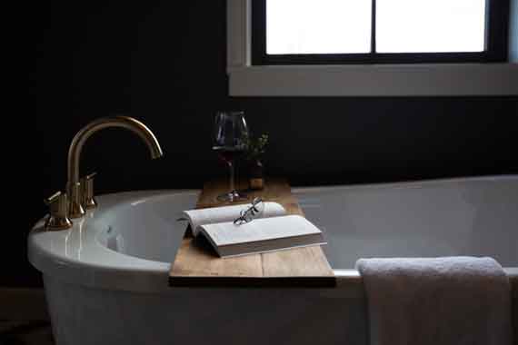 Open book and glasses on a wooden bath tray across a white freestanding tub, with a wine glass and towel in a dark bathroom.