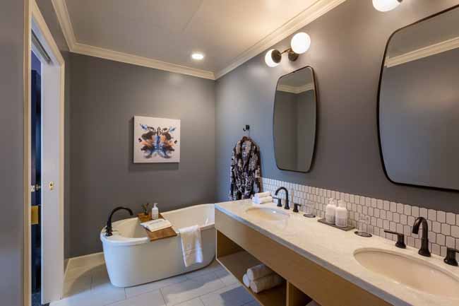 Modern bathroom with a double vanity, two oval mirrors, and a freestanding tub against gray walls; robe hangs by the sink area and artwork on the wall.