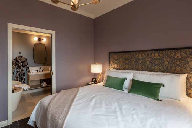 Hotel room with purple walls, a large bed with white linens and green pillows, and an open doorway to the en-suite bathroom showing a robe on a hook.