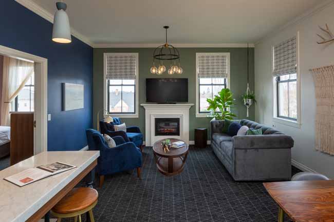 Cozy living room with a white fireplace mantel, mounted TV, and two windows letting in light; blue and sage walls with gray sofa and blue chairs.