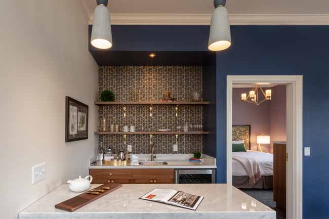 Compact kitchenette with a marble countertop, wood drawers, open shelves, and mosaic tile backsplash against a blue wall; doorway to a bedroom on the right.