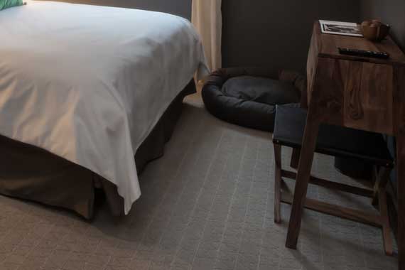 A bedroom corner with a made bed on the left, a black round pet bed on the floor, and a wooden desk with a stool on the right.