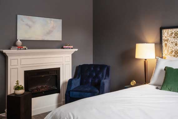 Cozy hotel-style bedroom corner with a white fireplace, dark gray walls, and a blue tufted chair by the mantle.