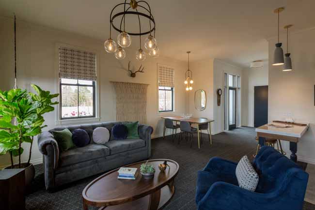 Bright open living room with a gray sofa, blue armchairs, a round wooden coffee table, a plant, and windows with woven shades; dining area visible in back.