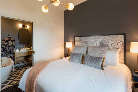 Hotel bedroom with a king-size bed, gray headboard, and two nightstands flanking lamps; open bathroom area visible to the left.