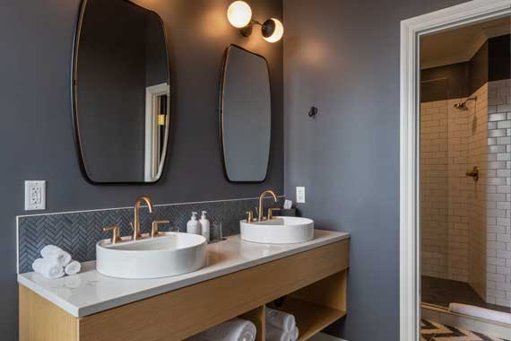 Double vanity with two white vessel sinks and gold faucets, two oval mirrors, gray walls, and a tiled backsplash; shelves with towels and a glass-tiled shower entrance to the right.
