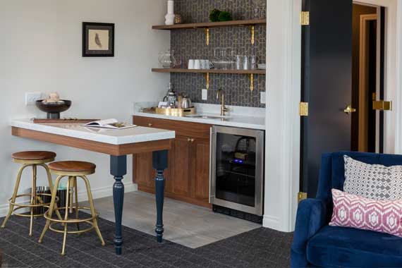 Modern living-room kitchenette with a white island, dark blue legs, two wooden stools, open shelves of glassware, and a stainless steel under-counter cooler; a blue sofa with pink pillows sits nearby.