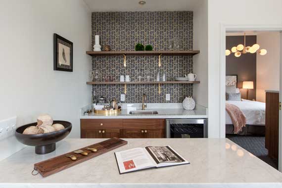Compact kitchen area with marble island, wooden shelves, and a tiled backsplash; doorway to a bedroom on the right.