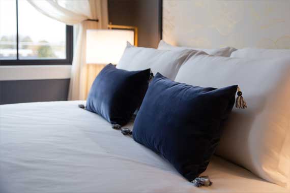 Luxurious hotel bed with white sheets and two navy blue throw pillows by a window.