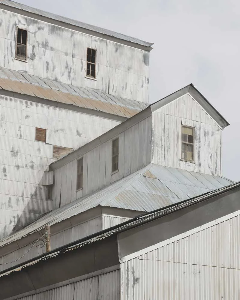 Weathered white metal warehouse with multiple gable roofs and small rectangular windows, showing peeling paint.