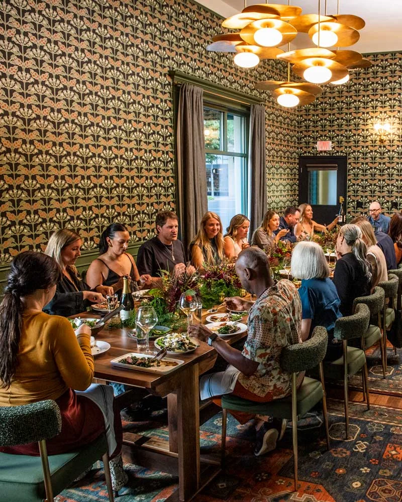 Atticus-Hotel-Meetings-Events A large group of people seated around a long wooden dining table in a warmly lit restaurant, sharing a meal with greenery centerpieces.