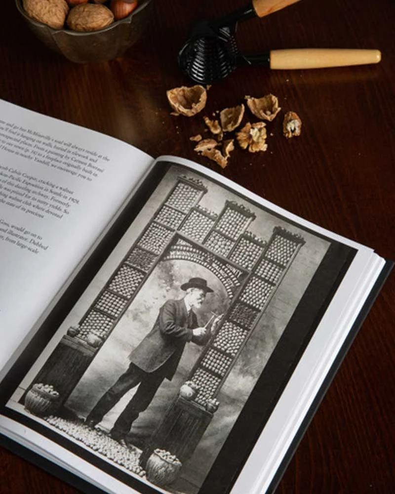 Open book displaying a vintage photo of a man in a suit and hat beside a lattice arch, with a nutcracker and walnut shells on the wooden table.
