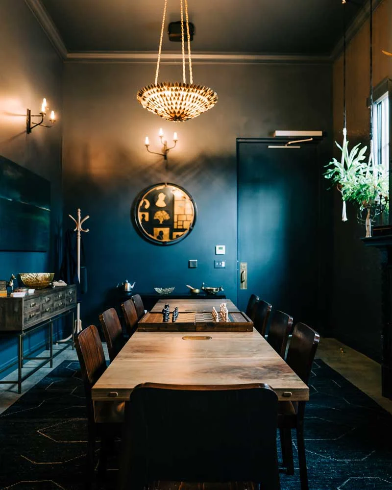 Long wooden dining table with a chessboard centerpiece in a dark blue-walled room, with a chandelier hanging above.