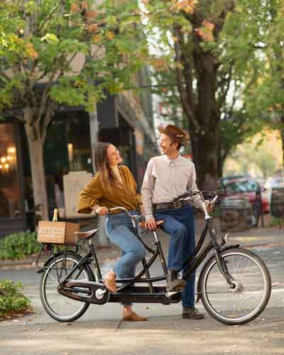 Two people ride a tandem bicycle on a tree-lined city street, smiling at each other.
